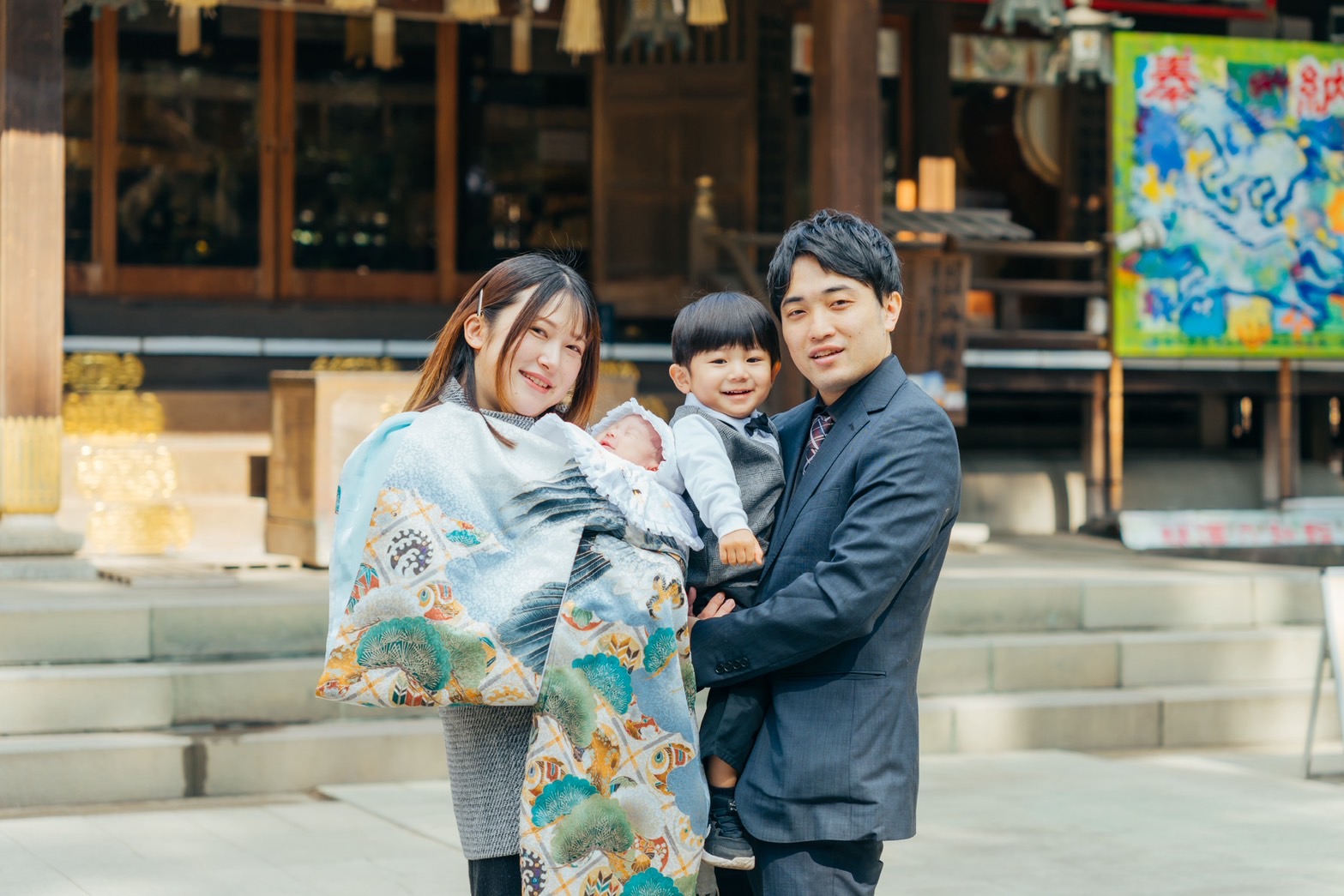 お宮参りで神社を訪れた家族写真。祝い着の赤ちゃんを抱くママと、スーツ姿のパパに抱かれて笑う男の子